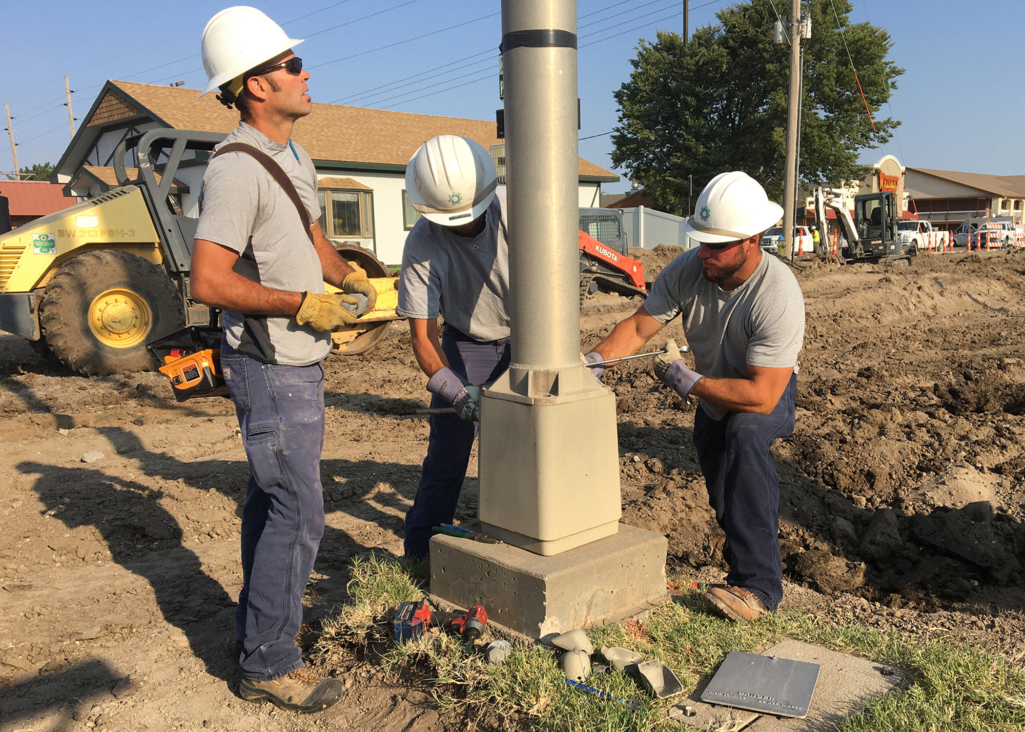 Three electric linemen use wrenches to detach a light pole from its base.  The workers are in grey shirts, wearing white helmets.