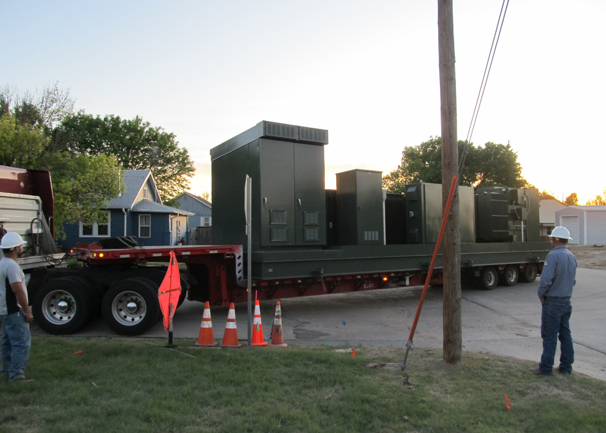 A skid-mounted substation, consisting of large green transformers and switch boxes, sits atop a flatbed semi trailer, waiting to be lifted into place.
