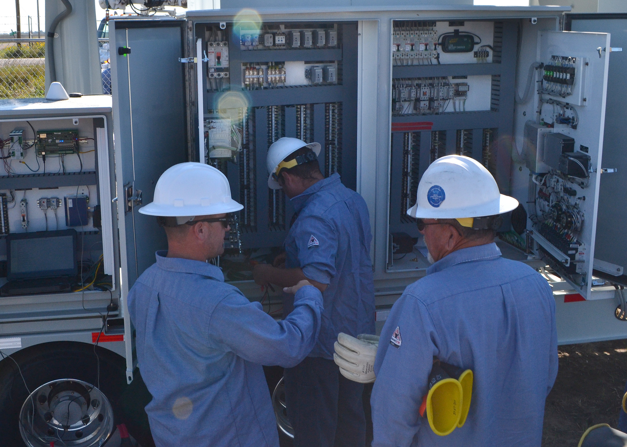 Three men in blue shirts and white hard hats talk as they look inside an electrical panel cabinet of a mobile substation.
