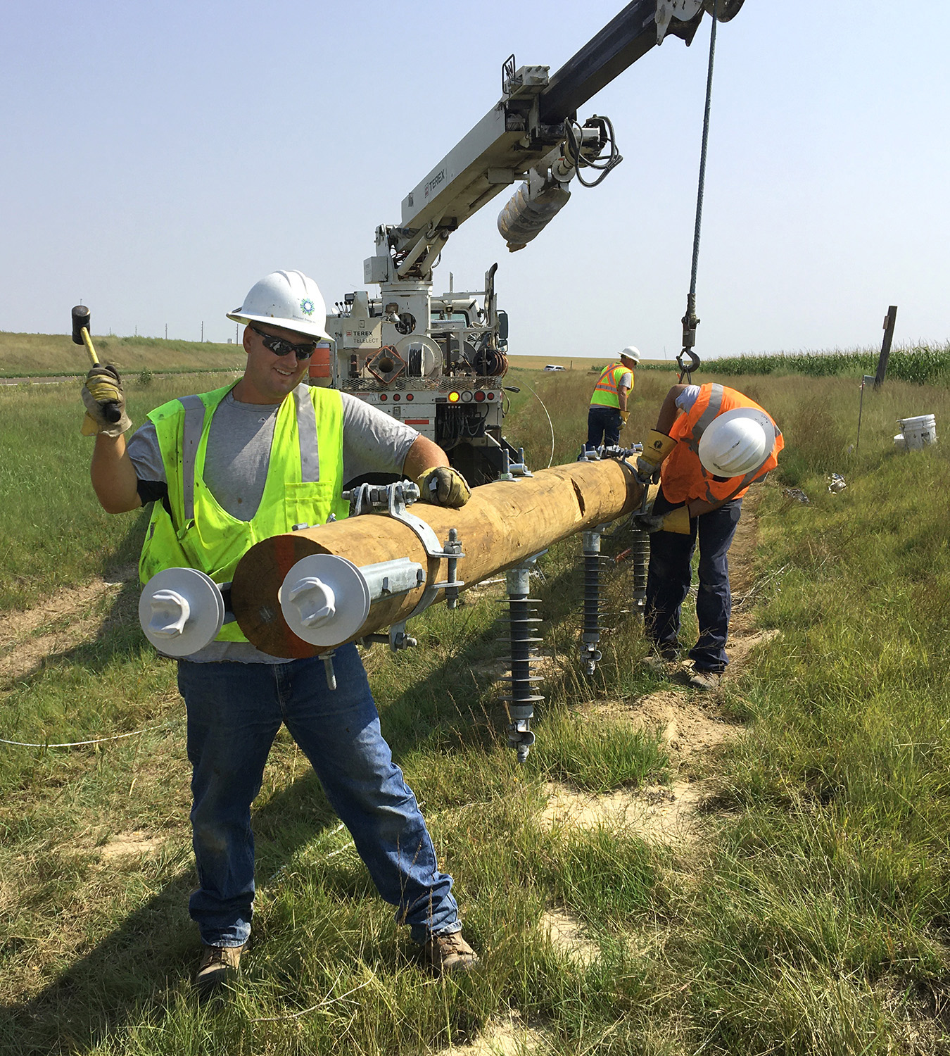 A lineman in a yellow vest hammers a steel brace into the top of power pole that is suspended near the ground by a truck's boom.