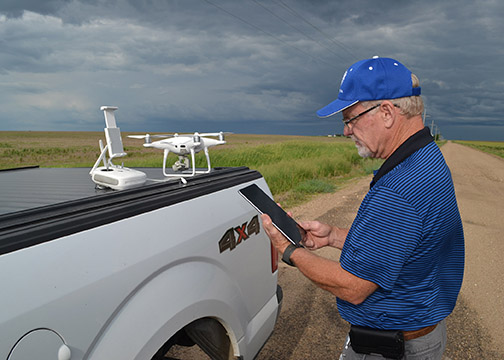 A man in a blue shirt stands at the bed of a pickup truck, on which a white drone sits.  The man is holding the drone's remote control, and is syncing it with the drone.