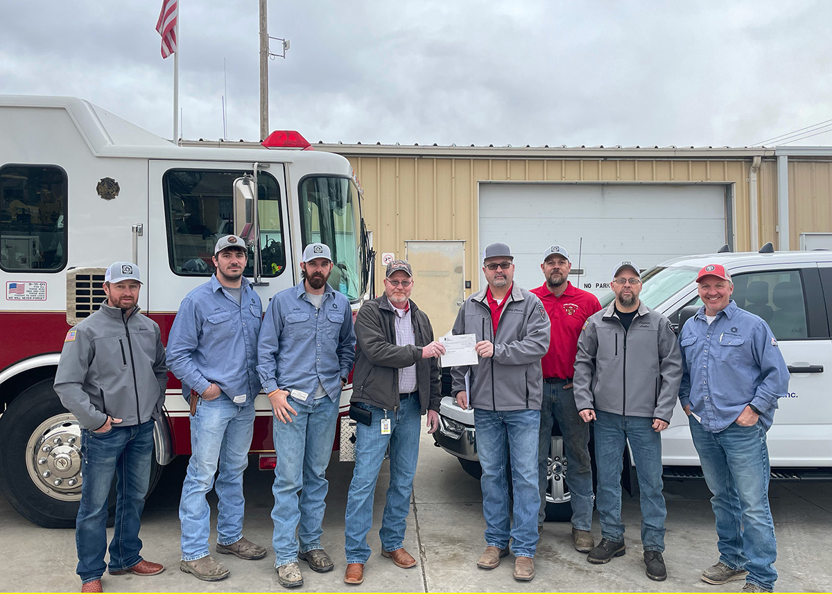 Eight men stand in front of a fire truck and a Midwest Energy vehicle. One Midwest Energy employee in the center hands a check to a member of the Phillipsburg Fire Department.