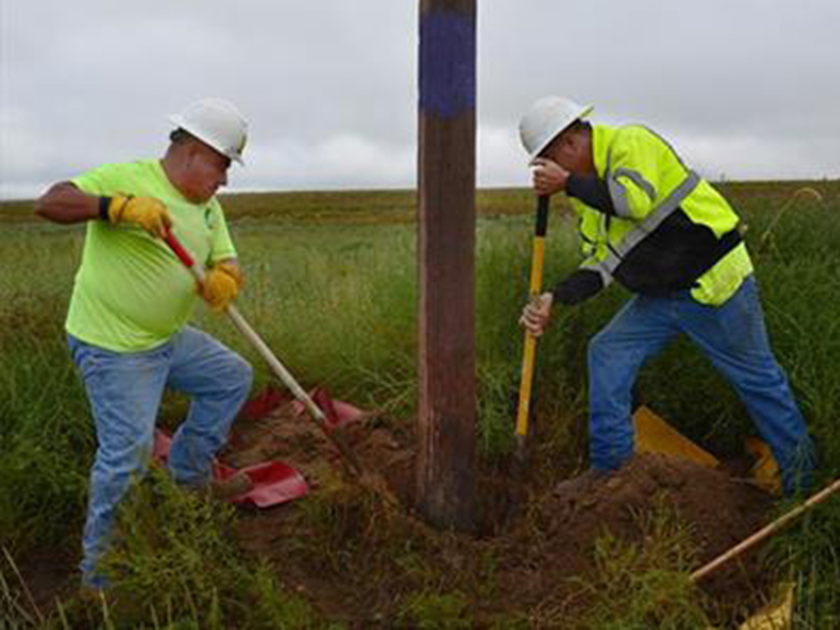 Two men in bright green safety shirts and hard hats shovel dirt from the base of a power pole to perform an inspection.