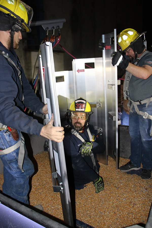 Three men stand in a grain bin. One is submerged in grain up to his waist. Two Larned firemen assemble a metal interlocking panels (a rescue grain tube) around him.