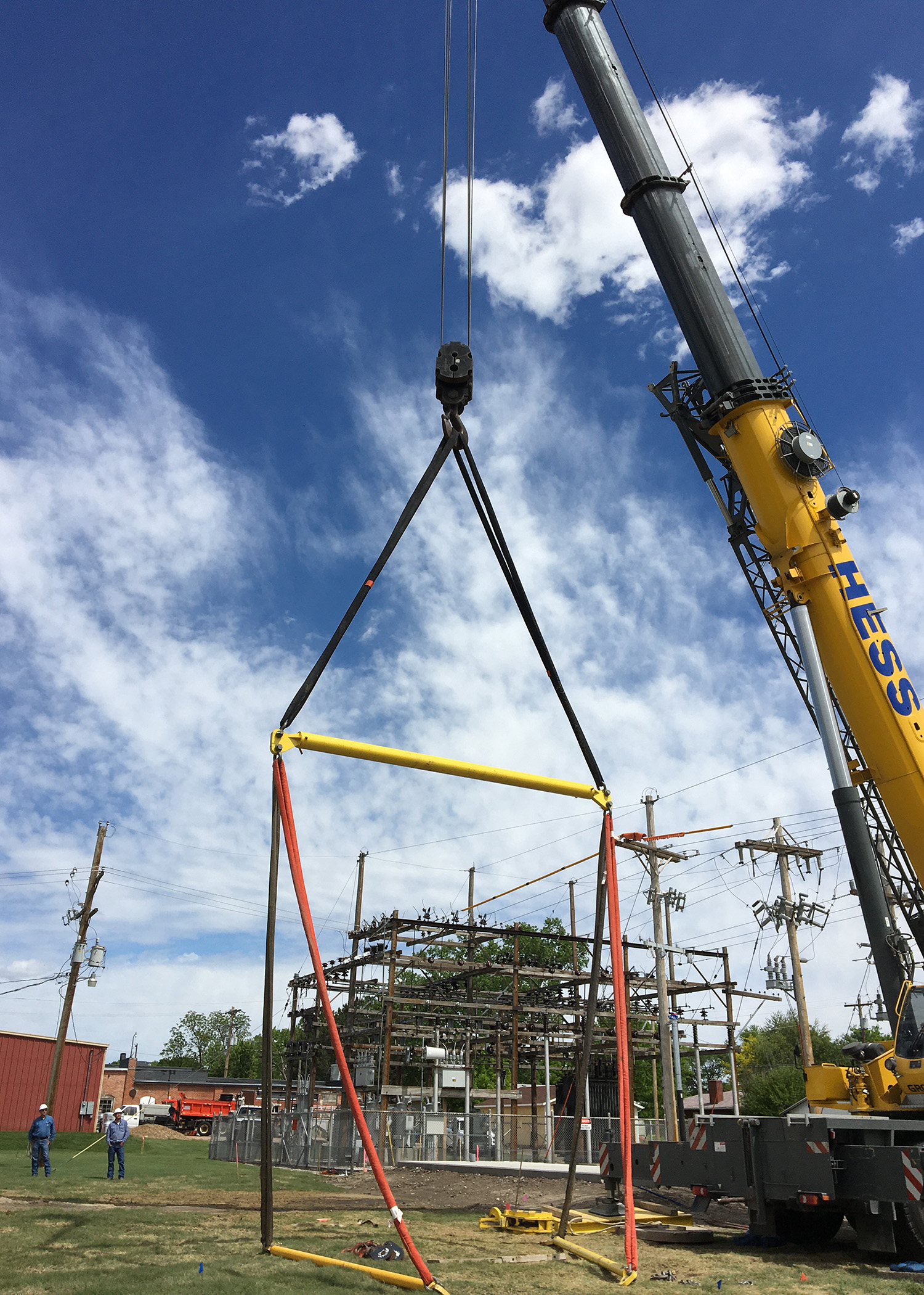 A very large crane from Hess Services is rigged with yellow harnesses to hoist a skid-mounted substation onto a concrete pad.