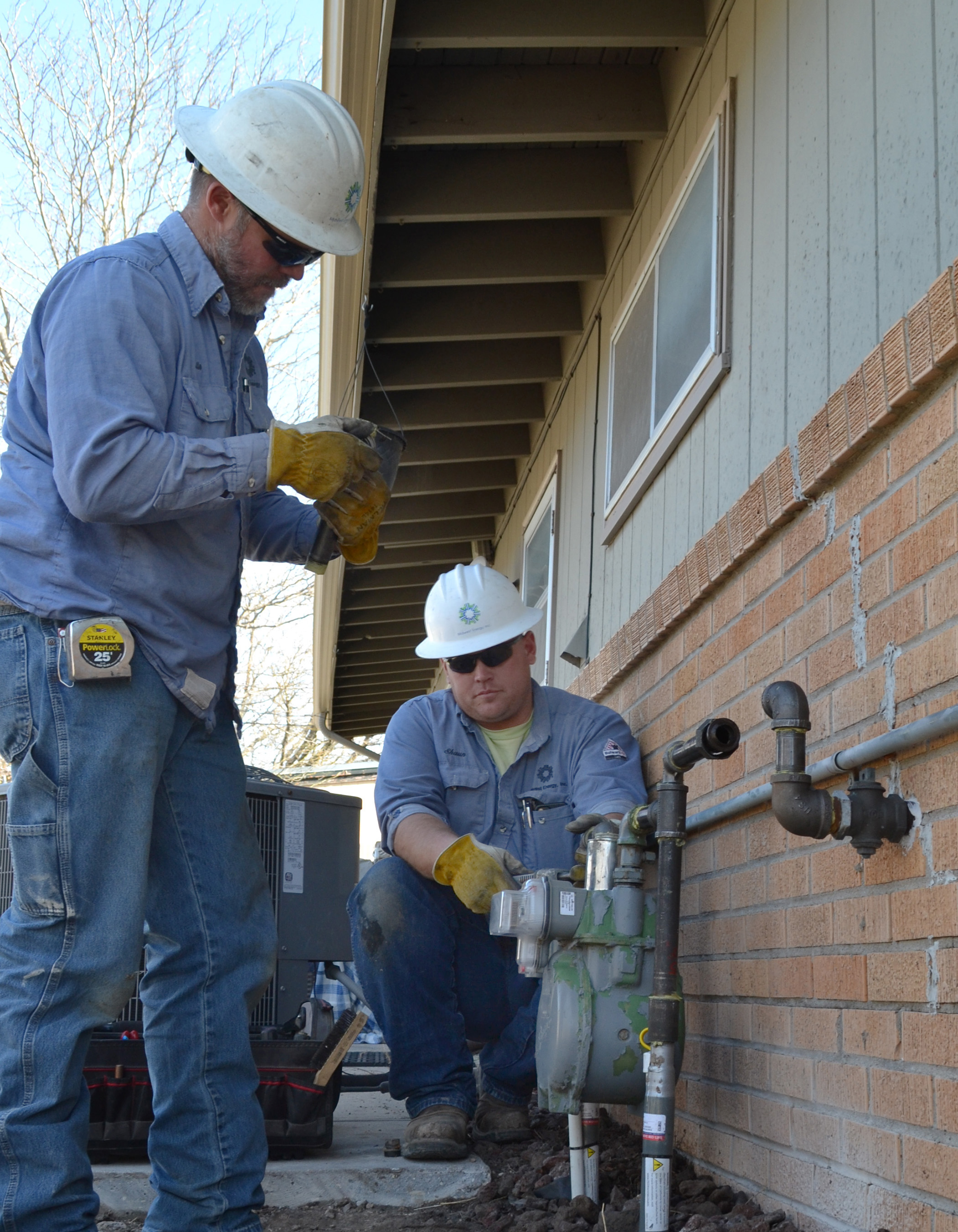 Two natural gas workers in blue shirts and white helmets attach a gas meter to the exterior of a brick home.
