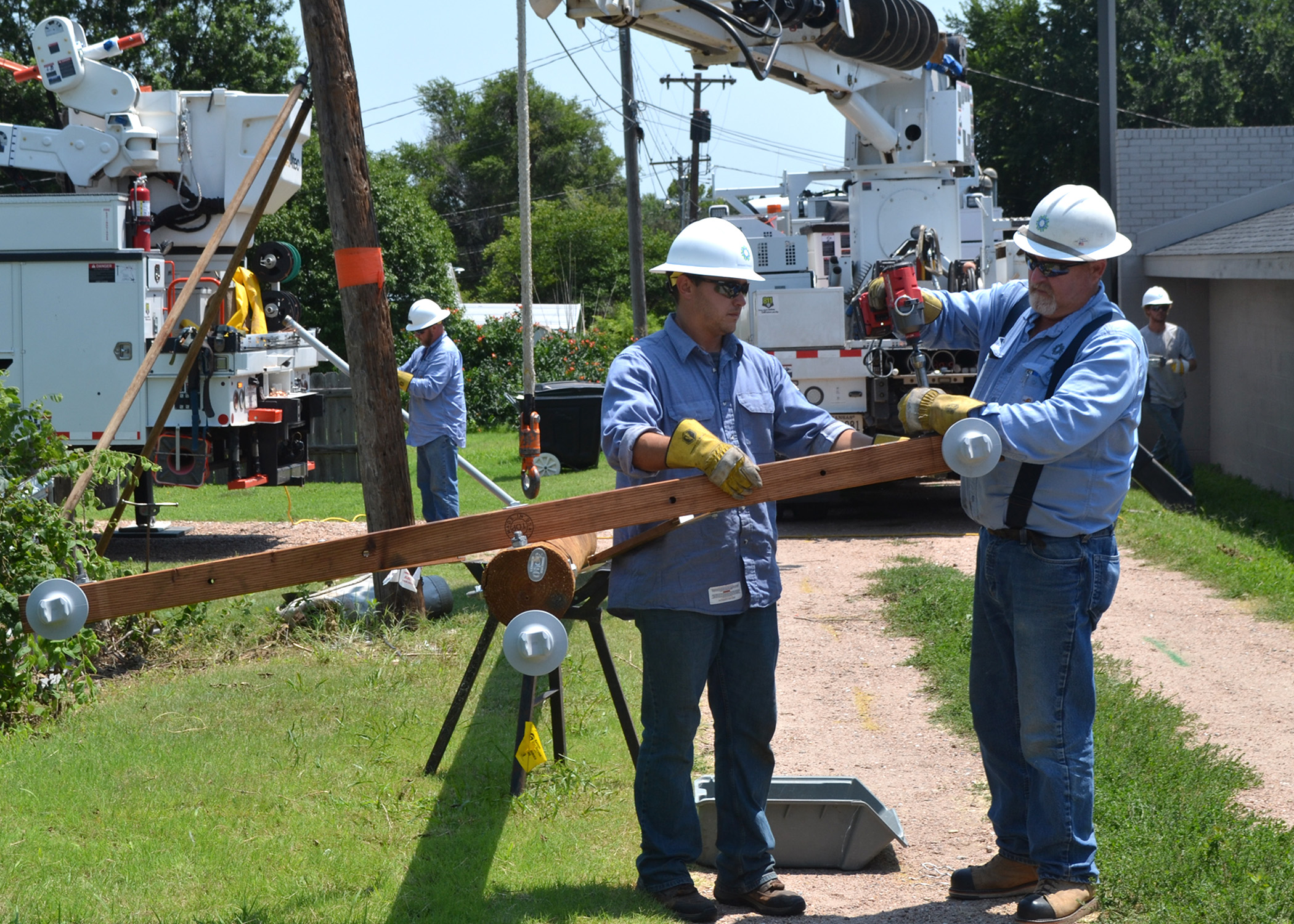 A photo of two electric linemen in blue shirts attaching hardware to a new pole that's on its side, in a narrow alleyway.  The pole is being suspended in the air by the boom from a white utility truck.