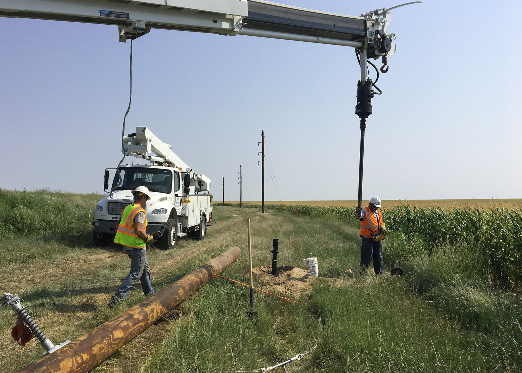 Two linemen in safety vests, working in a ditch near US-40, use a truck boom-mounted auger to drill a hole for a new power pole west of Winona, Kansas.