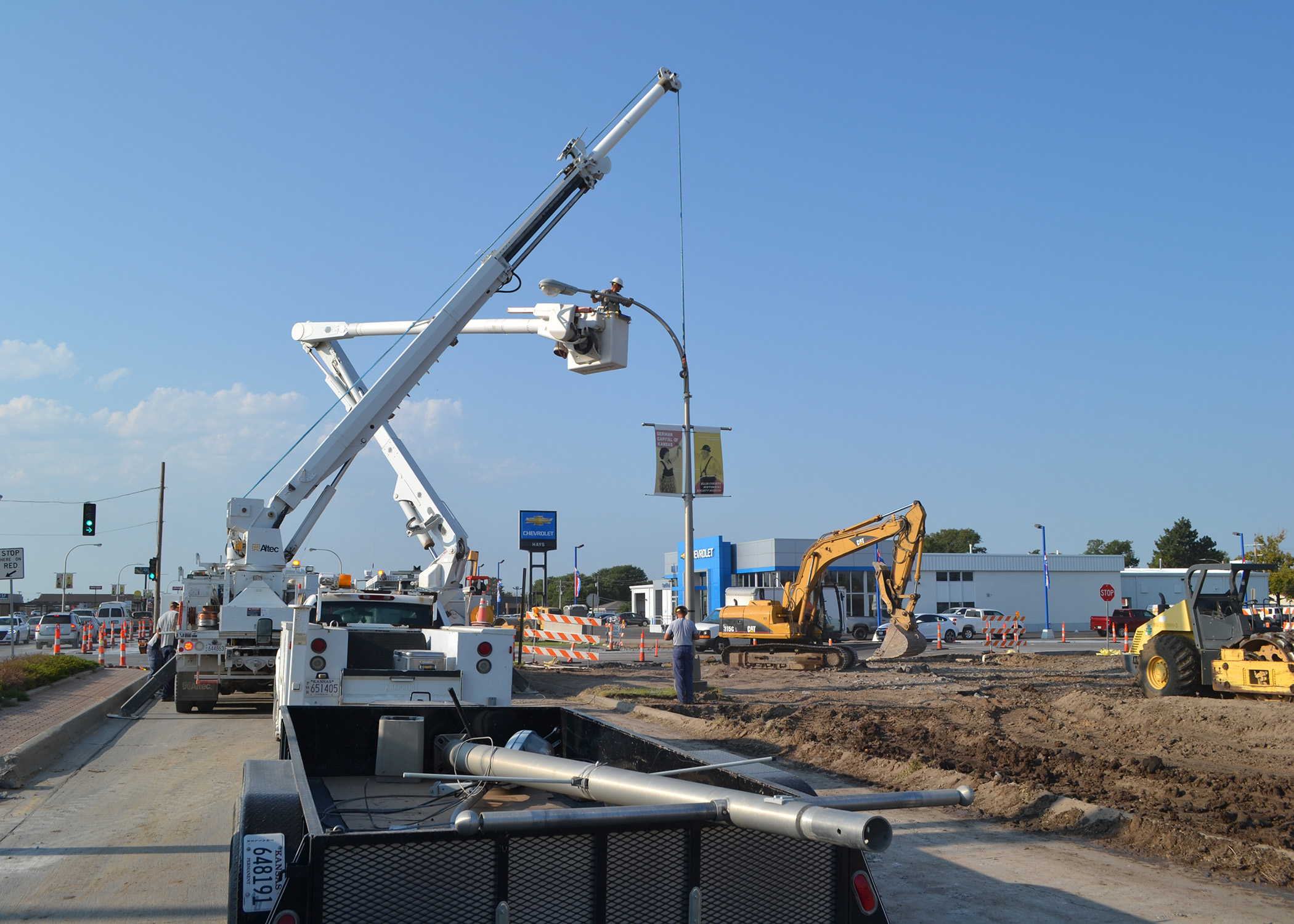 An image of a construction zone, with an electric crew truck lifting a streetlight off of its base; in the foreground are removed streetlights in a trailer, awaiting removal.