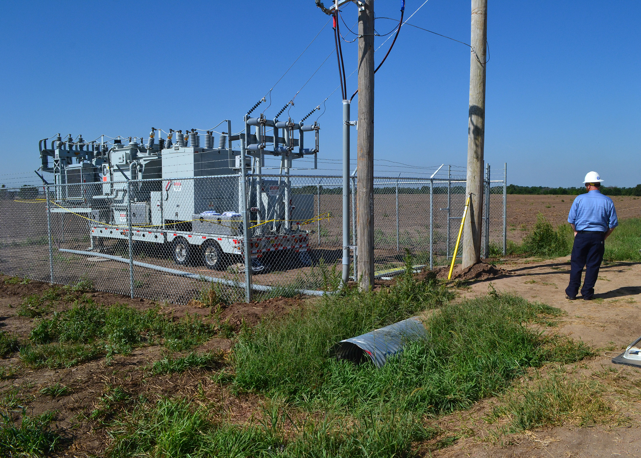 An image of a mobile electric substation, parked within a chain-link fence enclosure next to two power poles; a lineman in a blue shirt stands in a roadway and looks inward at the setup.
