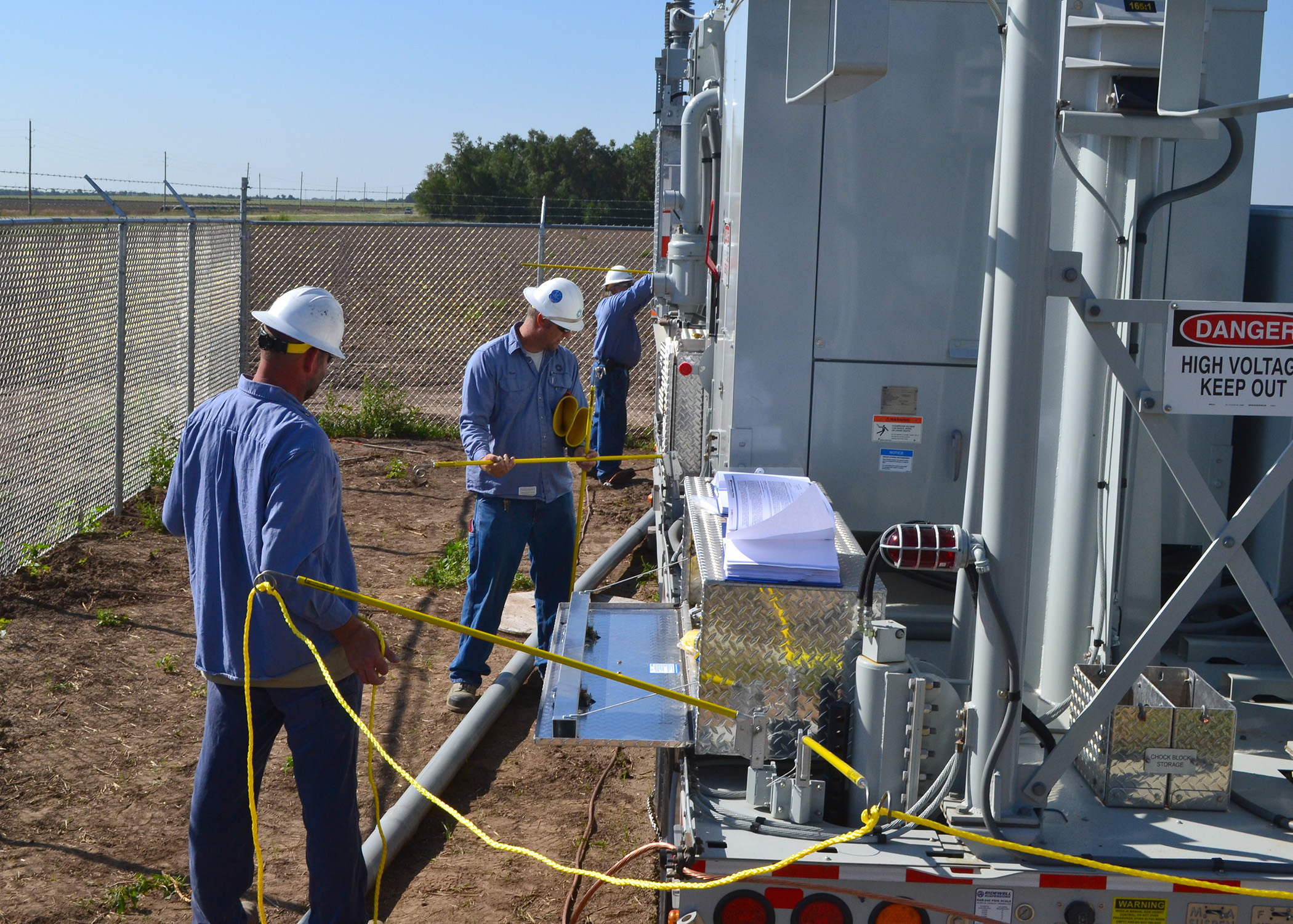 Three min in blue shirts and white hard hats install a yellow rope barrier around a mobile substation, designed to keep people from making contact with equipment once energized.