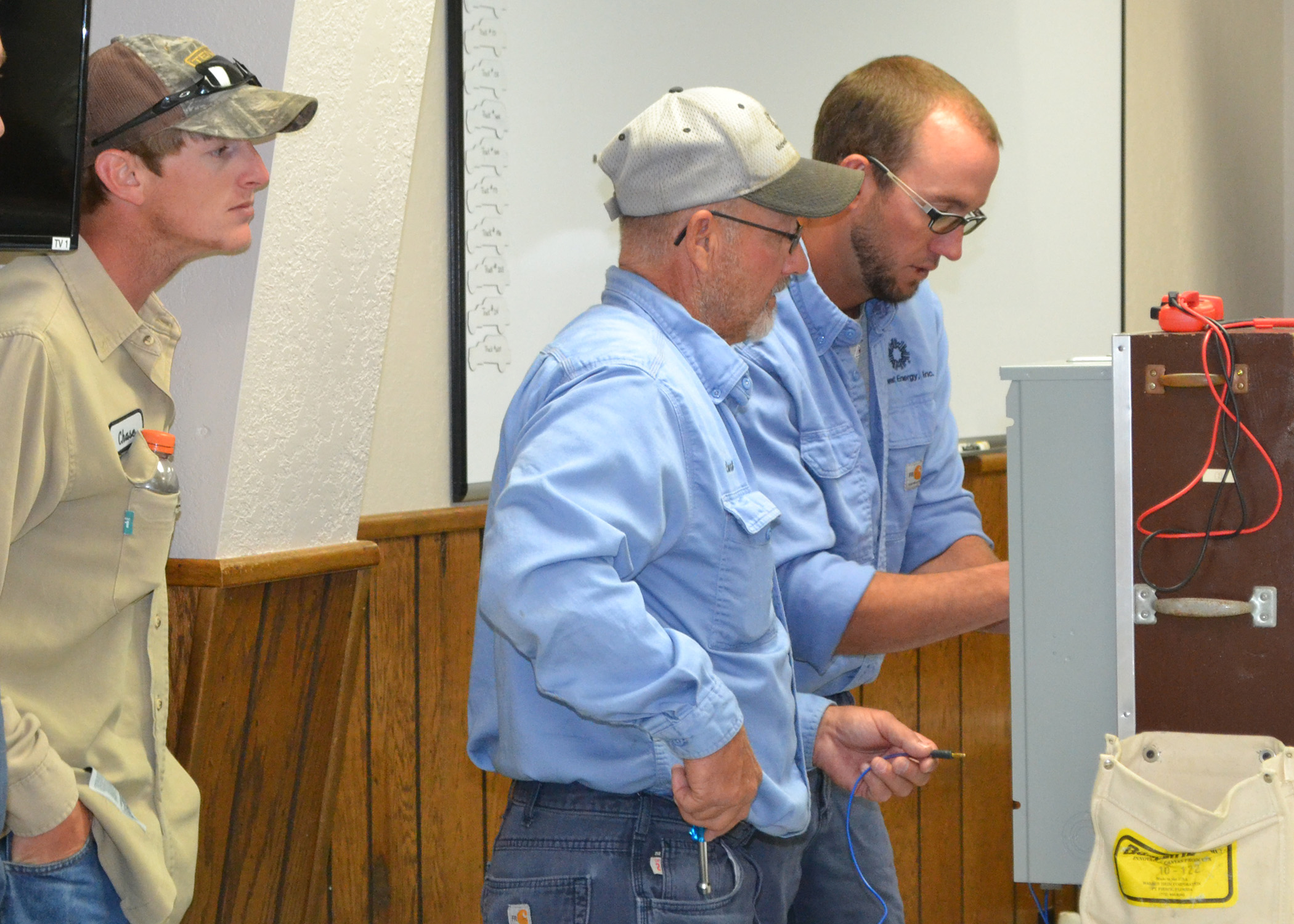 A picture of three linemen, one wearing a tan shirt and two wearing blue shirts, conducting a test on a meter can simulator during metering school.