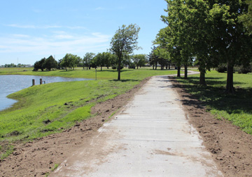 A new pedestrian and bicycle trail was partially paid for by a Midwest Energy grant. The photo shows the new concrete path with trees on the right side.  A slope to the left of the walkway leads to a lake.