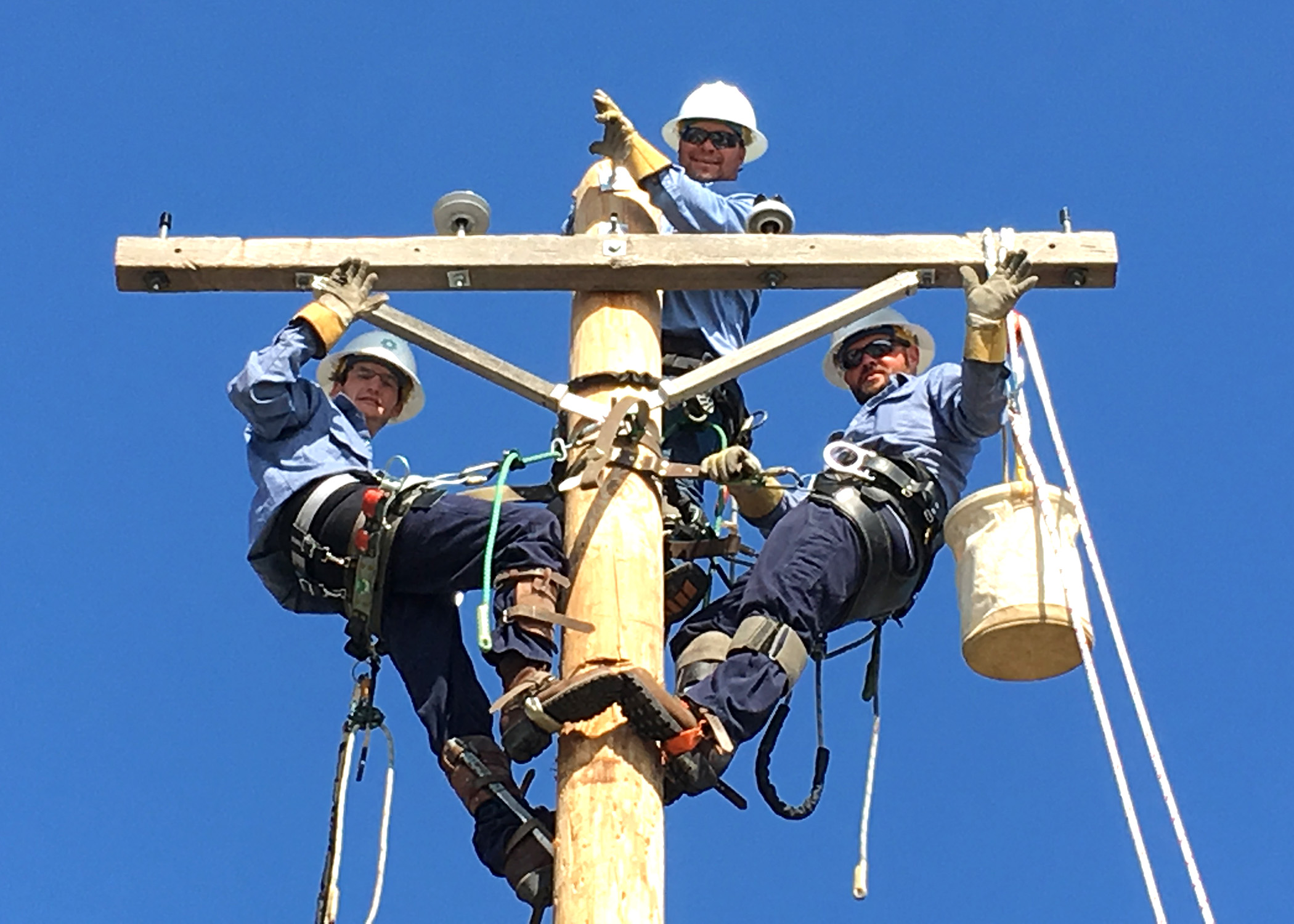 A photo of three linemen in blue shirts, manually climbing to the top of a training power pole, and looking down waving from atop the crossarm.