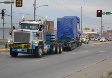 A natural gas generator, weighing 150 tons, is moved from the Hays Rail Yard up Vine St. in Hays on a special 130-foot trailer pulled by a white semi-truck. The generator is covered with a blue tarp.