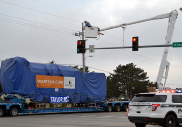 A natural gas generator, weighing 150 tons, is transported on Vine St. in Hays on a specialized trailer pulled by a white semi-truck. The generator is covered with a blue tarp. A Midwest Energy lineman, working from a bucket lift utility truck, raises a traffic signal to allow the generator to pass safely. A Hays Police Department SUV is in the lower right corner providing traffic control as the generator crosses the intersection.