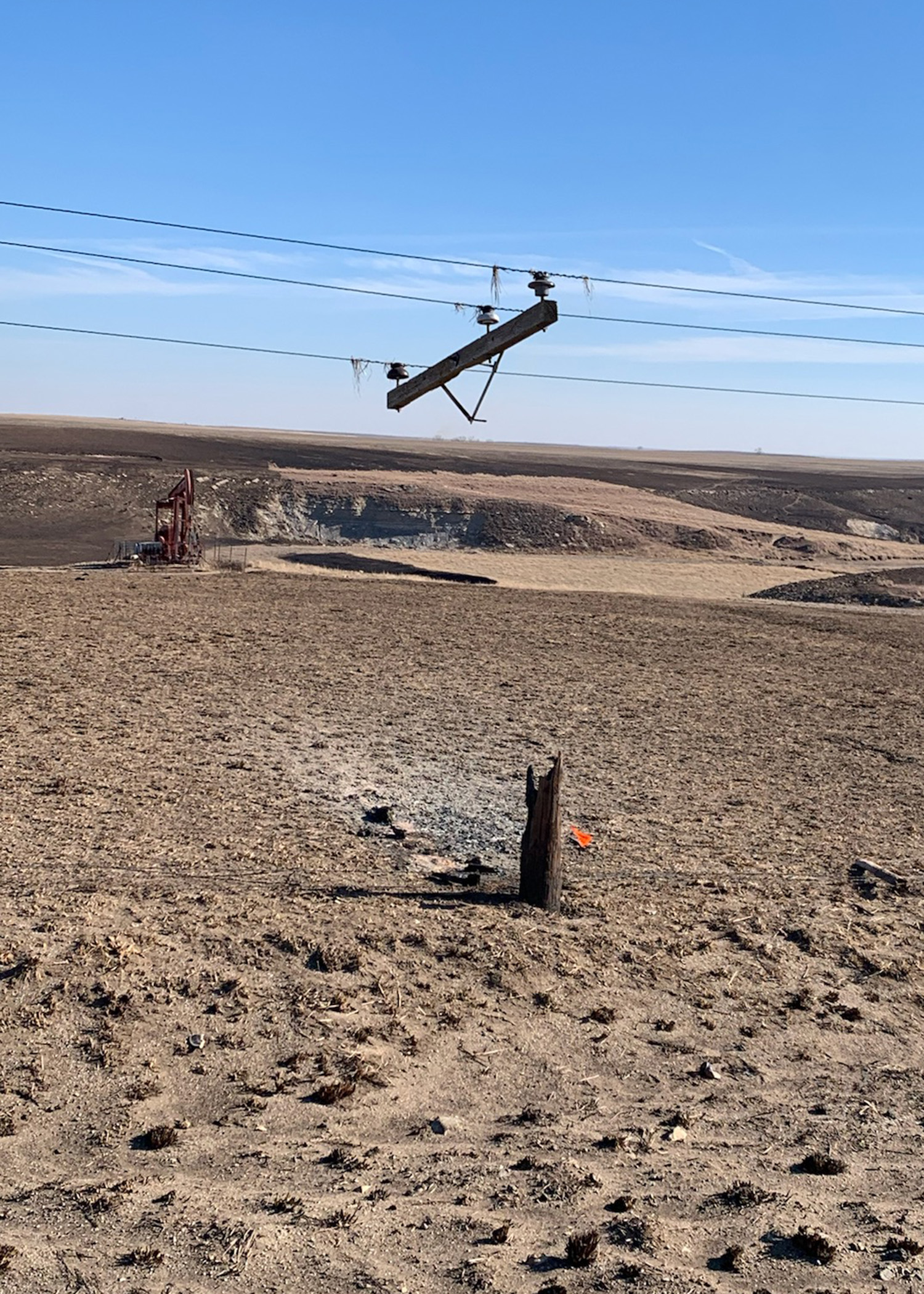 A power pole crossarm remains suspended in the air by the power lines, while the midsection of the power pole is burned away after a wildfire.