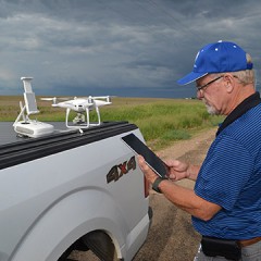 A man in a blue shirt stands at the bed of a pickup truck, on which a white drone sits.  The man is holding the drone's remote control, and is syncing it with the drone. 