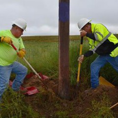 Two men in bright green safety shirts and hard hats shovel dirt from the base of a power pole to perform an inspection.  