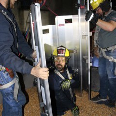 Three men stand in a grain bin. One is submerged in grain up to his waist. Two Larned firemen assemble a metal interlocking panels (a rescue grain tube) around him. 