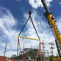 A very large crane from Hess Services is rigged with yellow harnesses to hoist a skid-mounted substation onto a concrete pad.  