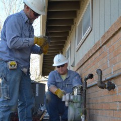 Two natural gas workers in blue shirts and white helmets attach a gas meter to the exterior of a brick home.  