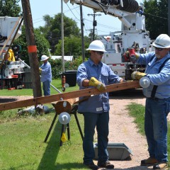 A photo of two electric linemen in blue shirts attaching hardware to a new pole that's on its side, in a narrow alleyway.  The pole is being suspended in the air by the boom from a white utility truck.