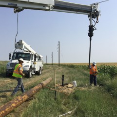 Two linemen in safety vests, working in a ditch near US-40, use a truck boom-mounted auger to drill a hole for a new power pole west of Winona, Kansas.  