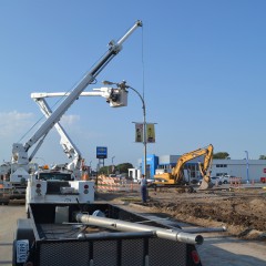 An image of a construction zone, with an electric crew truck lifting a streetlight off of its base; in the foreground are removed streetlights in a trailer, awaiting removal.