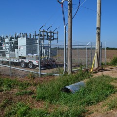 An image of a mobile electric substation, parked within a chain-link fence enclosure next to two power poles; a lineman in a blue shirt stands in a roadway and looks inward at the setup.  