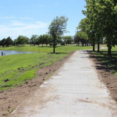 A new pedestrian and bicycle trail was partially paid for by a Midwest Energy grant. The photo shows the new concrete path with trees on the right side.  A slope to the left of the walkway leads to a lake.