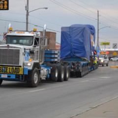 A natural gas generator, weighing 150 tons, is moved from the Hays Rail Yard up Vine St. in Hays on a special 130-foot trailer pulled by a white semi-truck. The generator is covered with a blue tarp.