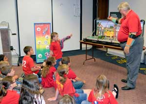 A man in a red shirt uses a tabletop display to demonstrate electric safety to a room full of young children, also in red shirts.